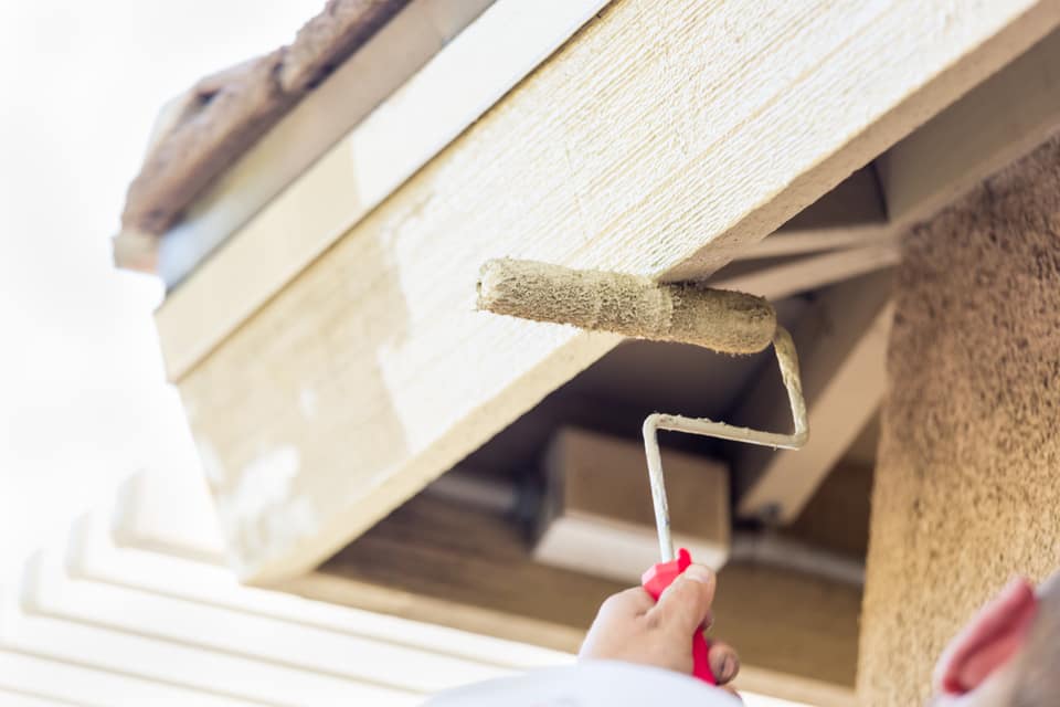 Painter rolling fresh paint onto the underside of an exterior roof overhang and siding