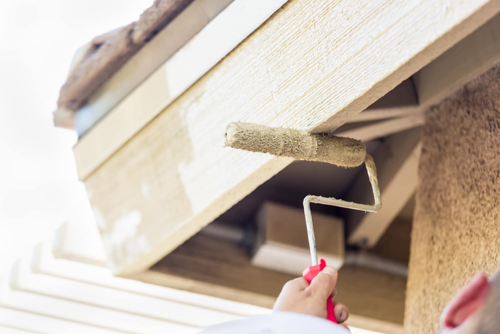 Painter rolling fresh paint onto the underside of an exterior roof overhang and siding
