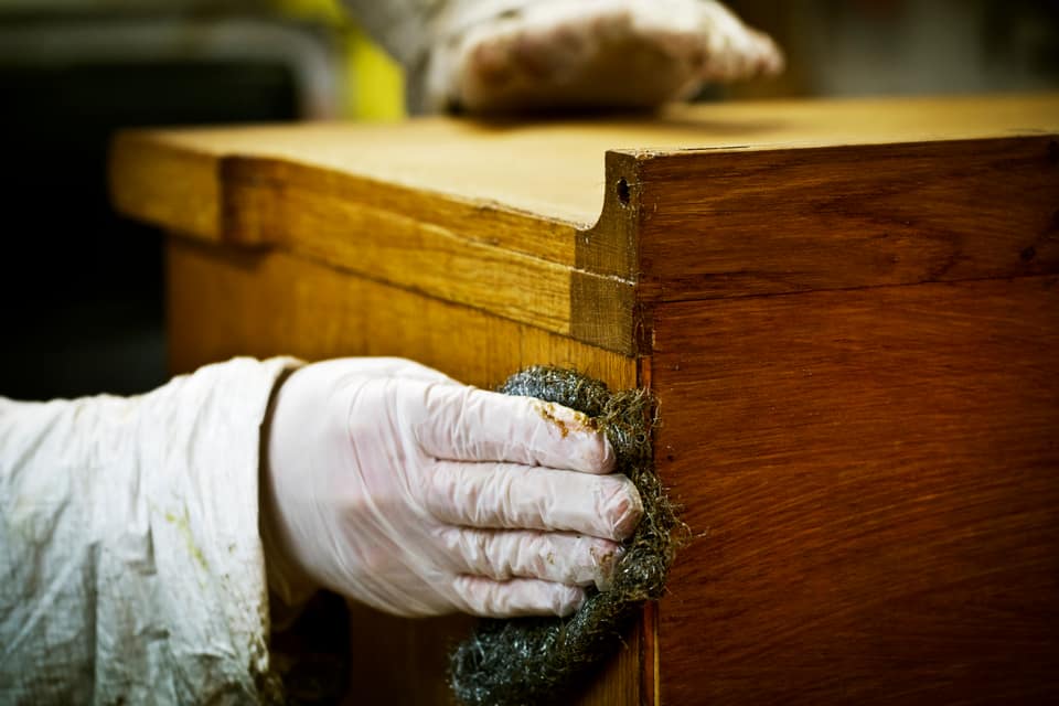 Gloved hand sanding and scouring a wooden surface back to bare timber before finishing